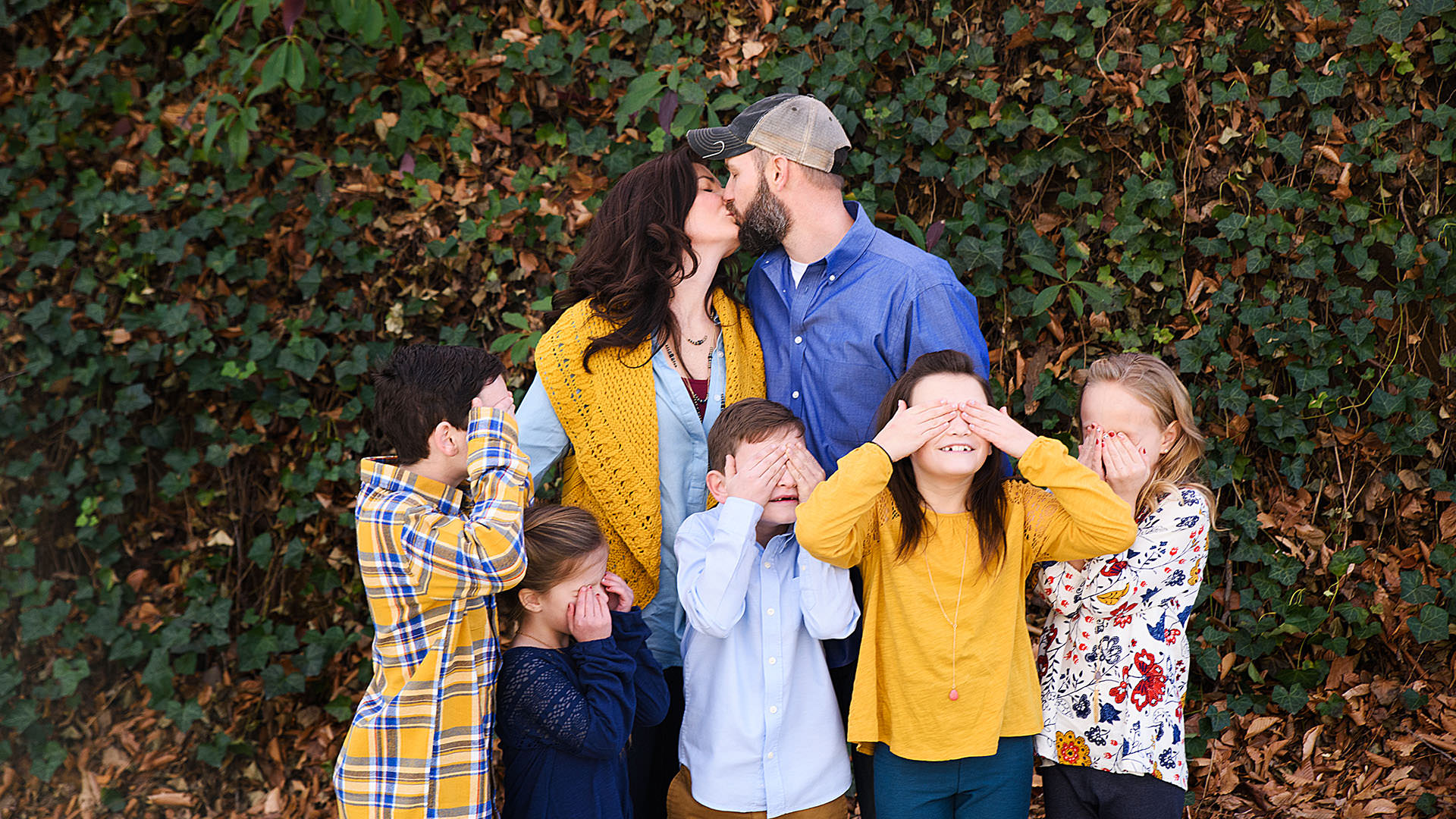 Creative shot of a family where the mom and dad are kissing while the children are holding their eyes shut.